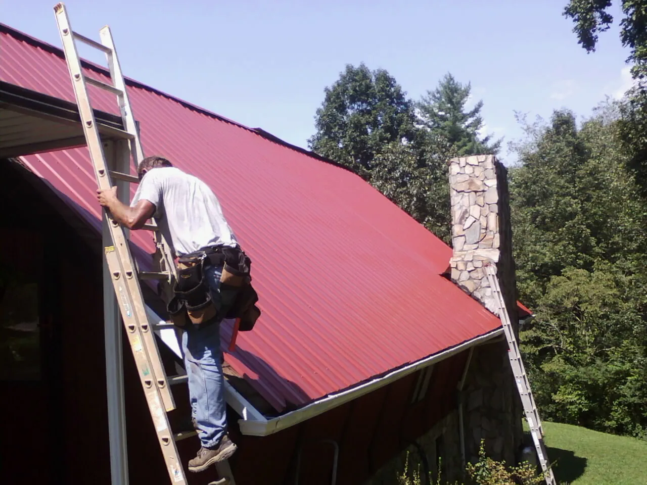 Roofer on a ladder beside a red metal roof and stone chimney — Soffit & Fascia Repair by Roofing Brothers in Gaithersburg